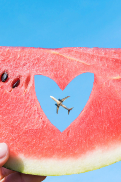 Ripe piece of watermelon with heart shape hole in female hands on the background of the blue sky and plane.